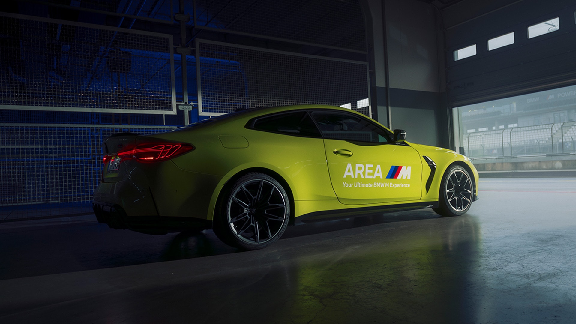 A bright yellow BMW M sports car parked inside what appears to be a garage or workshop with blue lighting. The car has the text 'AREA M Your Ultimate BMW M Experience' printed on its side door. The vehicle features distinctive red taillights and black alloy wheels, positioned on a polished concrete floor with large windows visible in the background.