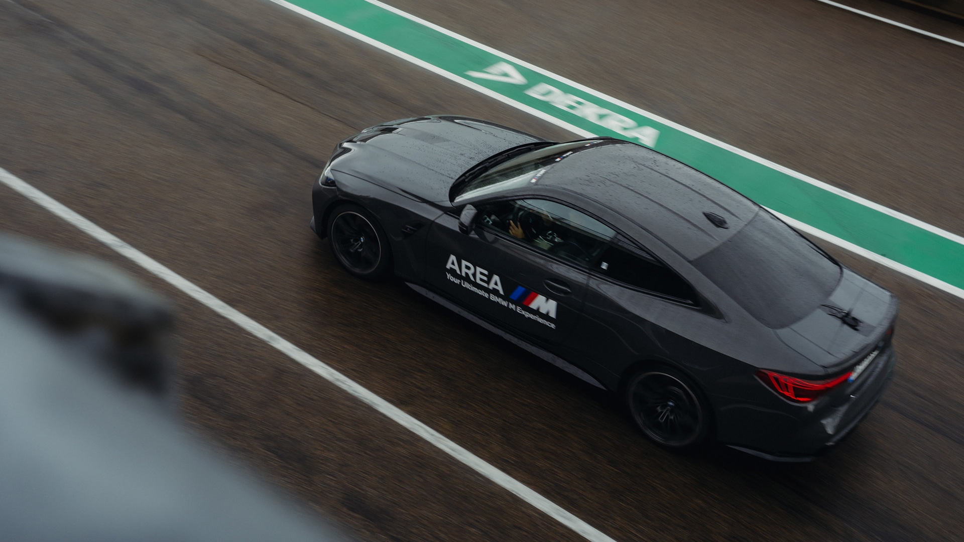 A black BMW M series sports car driving on a racetrack with a green pit lane stripe visible. The car has 'AREA M' branding on its side and is photographed from above in motion.