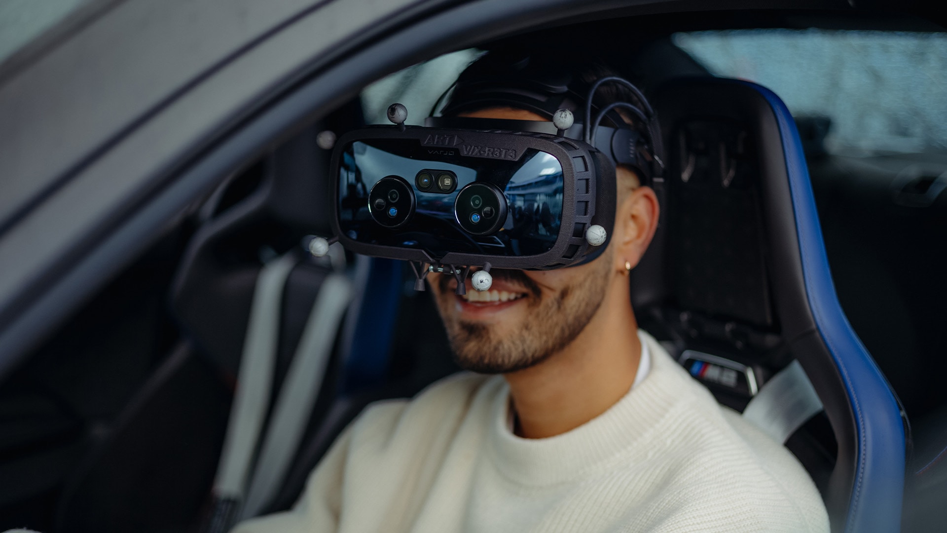A person sitting in a car with blue interior seats wearing a virtual reality headset. The person is dressed in a white sweater and appears to be smiling while using the VR device, which has multiple sensors or cameras visible on its front.