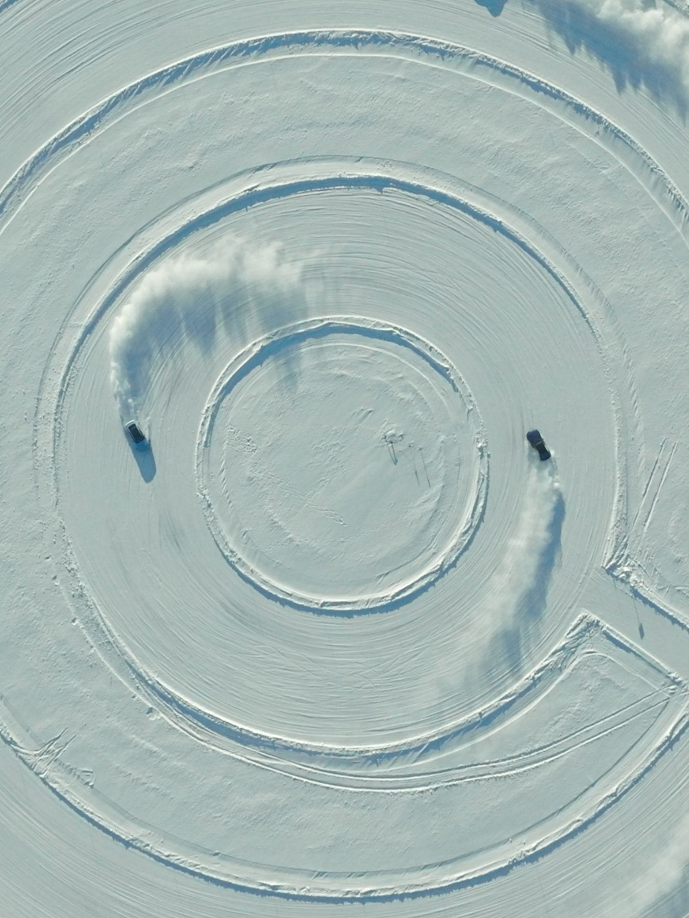 Aerial view of two vehicles driving on a circular snow track, creating spiral patterns in the white snow. The vehicles are leaving trails as they navigate around the concentric circular paths carved into the snowy surface.