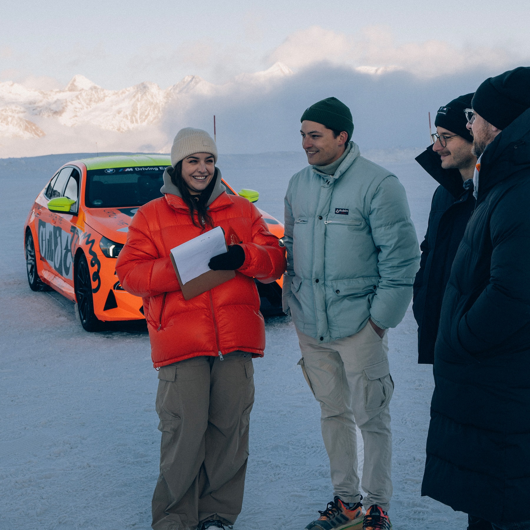 A group of people in winter clothing standing on a snowy landscape with mountains in the background. A bright orange sports car with neon green accents is parked behind them. One person in a red puffer jacket is holding papers while conversing with others wearing light and dark winter coats and beanies.