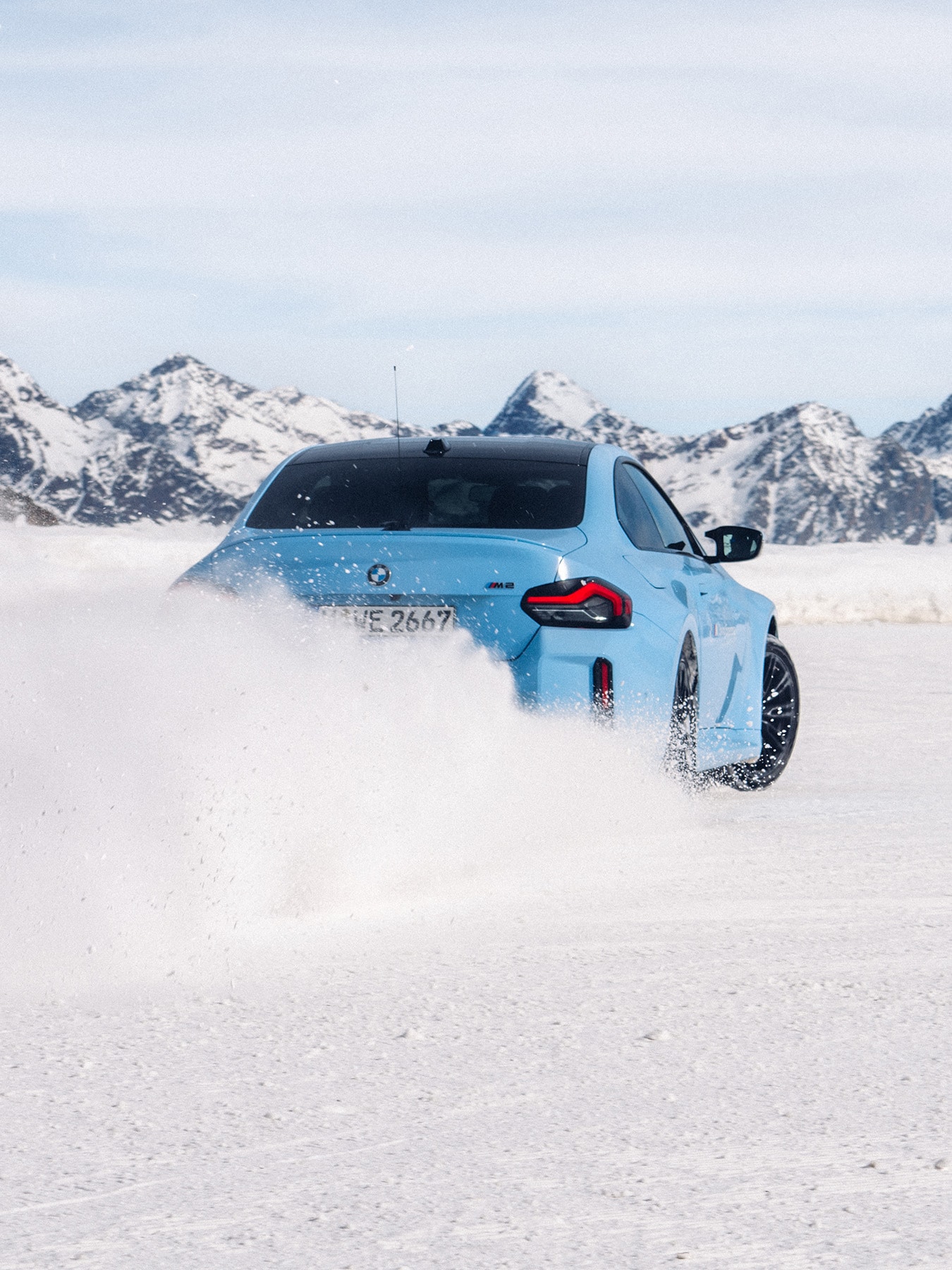 A light blue BMW sports car driving through snow, kicking up a spray of powder as it moves across a snowy landscape. Snow-capped mountains rise in the background against a pale blue sky.