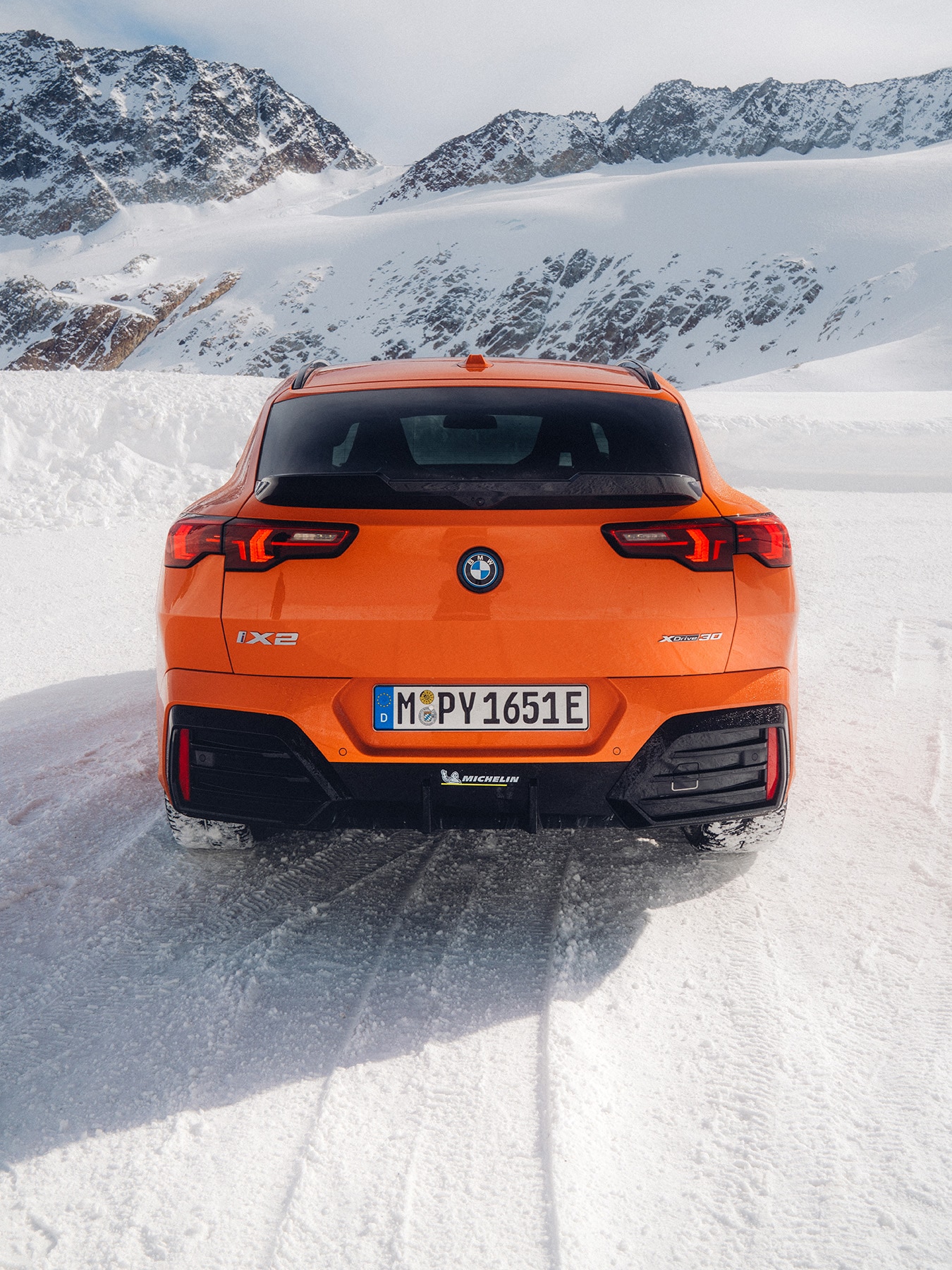 Rear view of an orange BMW iX2 xDrive30 electric vehicle parked on a snow-covered landscape with snow-capped mountains in the background. The car displays a German license plate and Michelin branding.