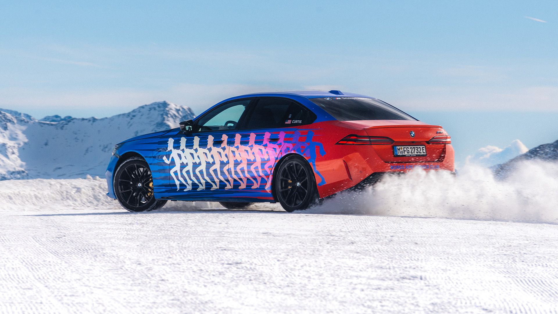 A BMW sedan with a distinctive blue and red color scheme featuring white silhouette figures in a running motion across the side. The car is driving on snow, kicking up powder, with snow-covered mountains in the background under a clear blue sky.