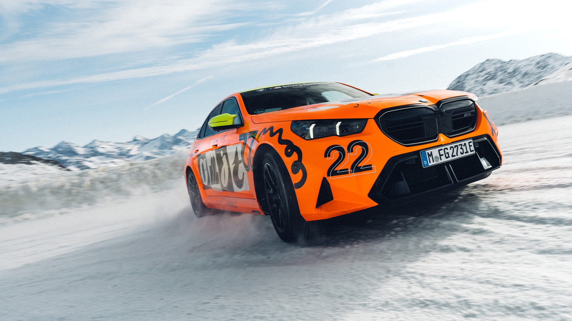 A bright orange sports car with the number 22 prominently displayed on its side drifts across a snowy landscape. The vehicle, which appears to be a BMW with a German license plate, kicks up snow as it navigates through a winter mountain setting with snow-capped peaks visible in the background under a blue sky with wispy clouds.