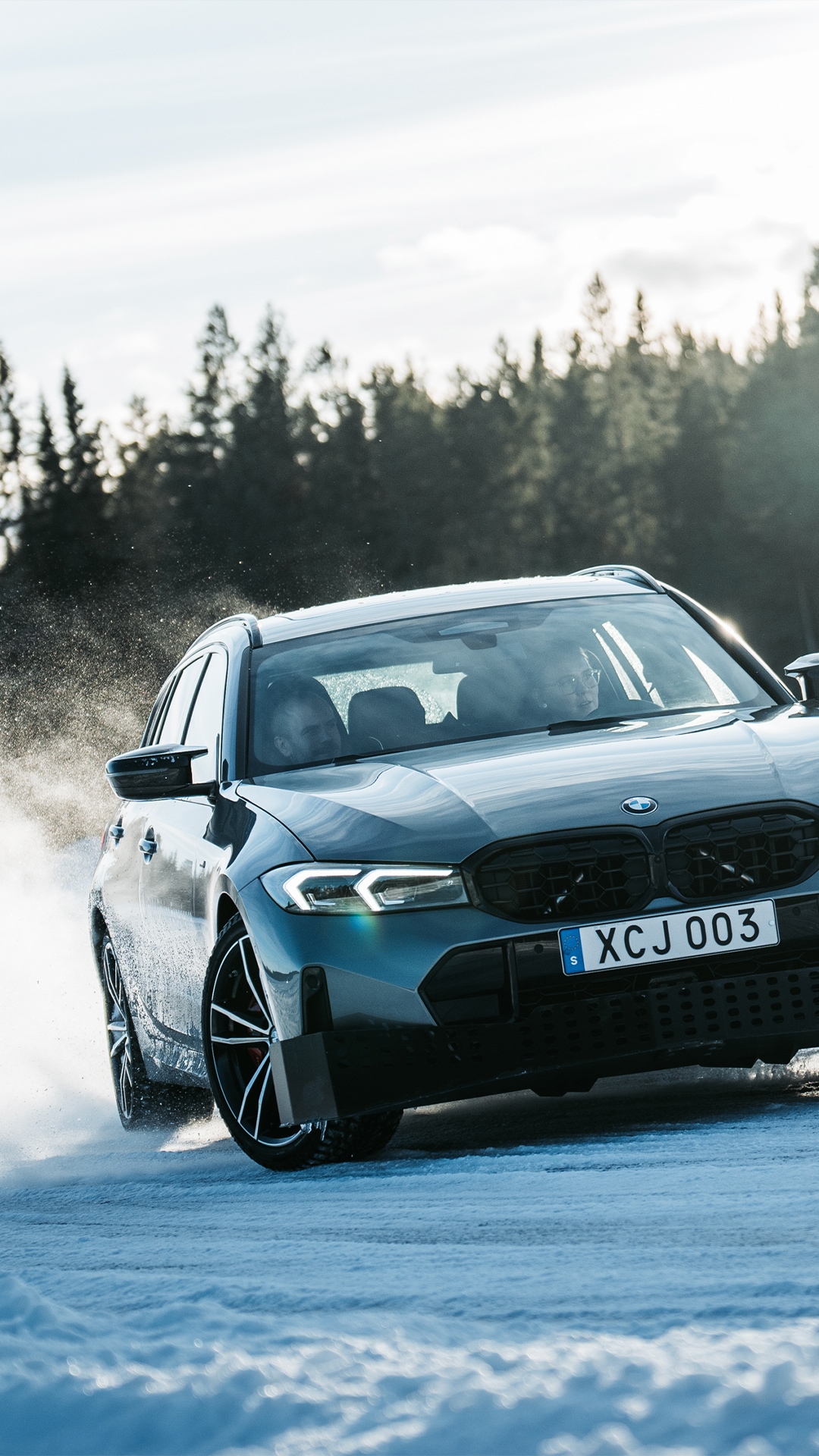 A dark blue BMW driving on a snowy surface, kicking up snow spray as it moves. The car has a Swedish license plate and is set against a backdrop of evergreen trees under an overcast sky.