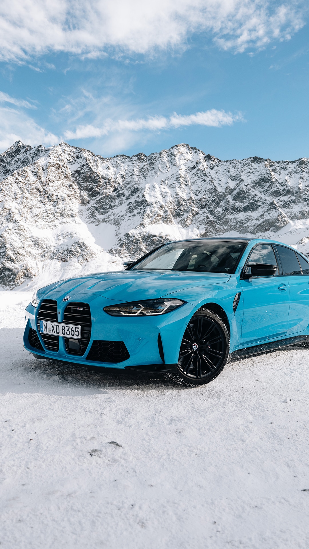 A bright blue BMW sports car parked on snow-covered ground against a backdrop of snow-capped mountains. The vehicle features distinctive black wheels and the large BMW kidney grille. Above, a blue sky with scattered white clouds completes the alpine winter scene.