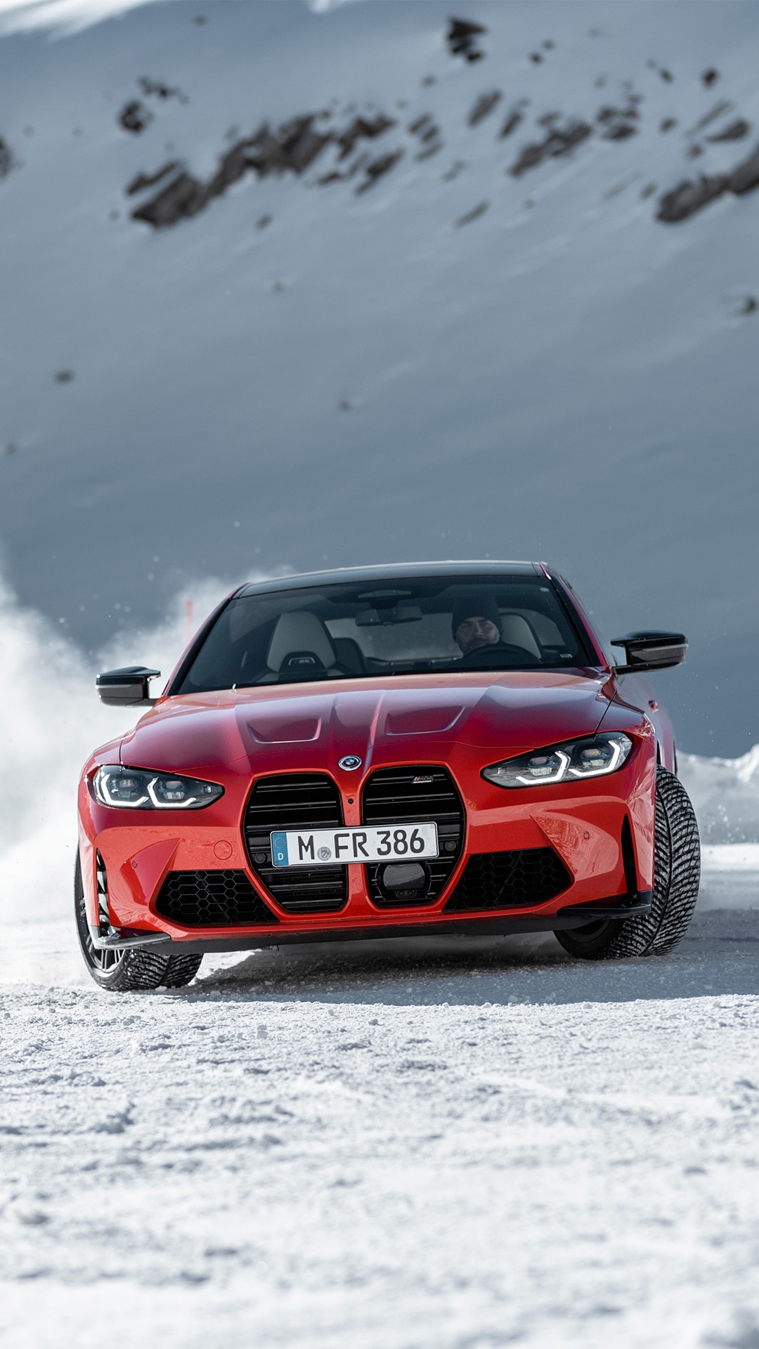 A bright red BMW sports car with German license plate driving on a snowy surface. The vehicle features the distinctive large kidney grille and is photographed head-on against a mountainous winter backdrop.