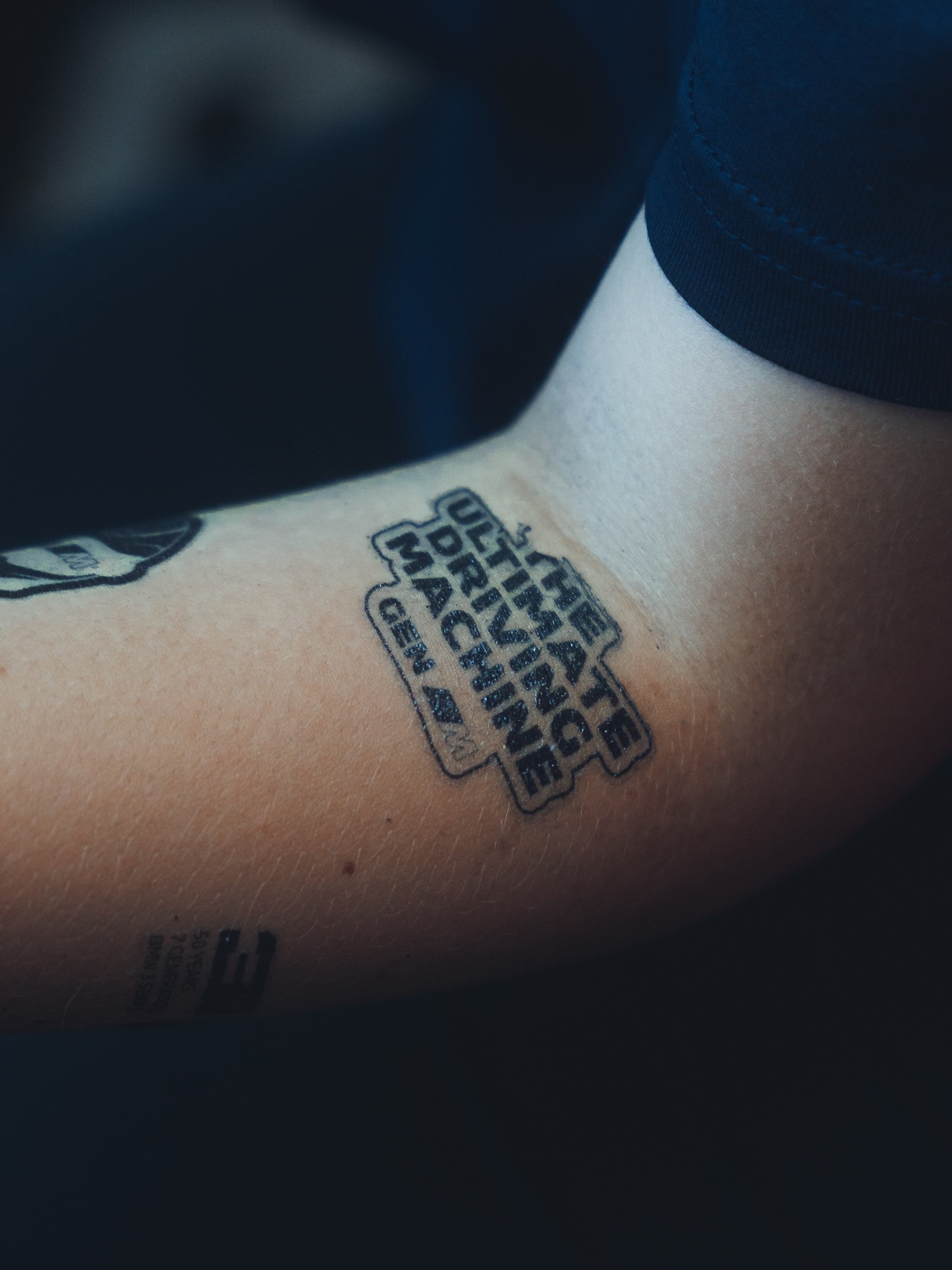 A Gen-M-Tatoo on a young man's forearm saying "The Ultimative Driving Machine".