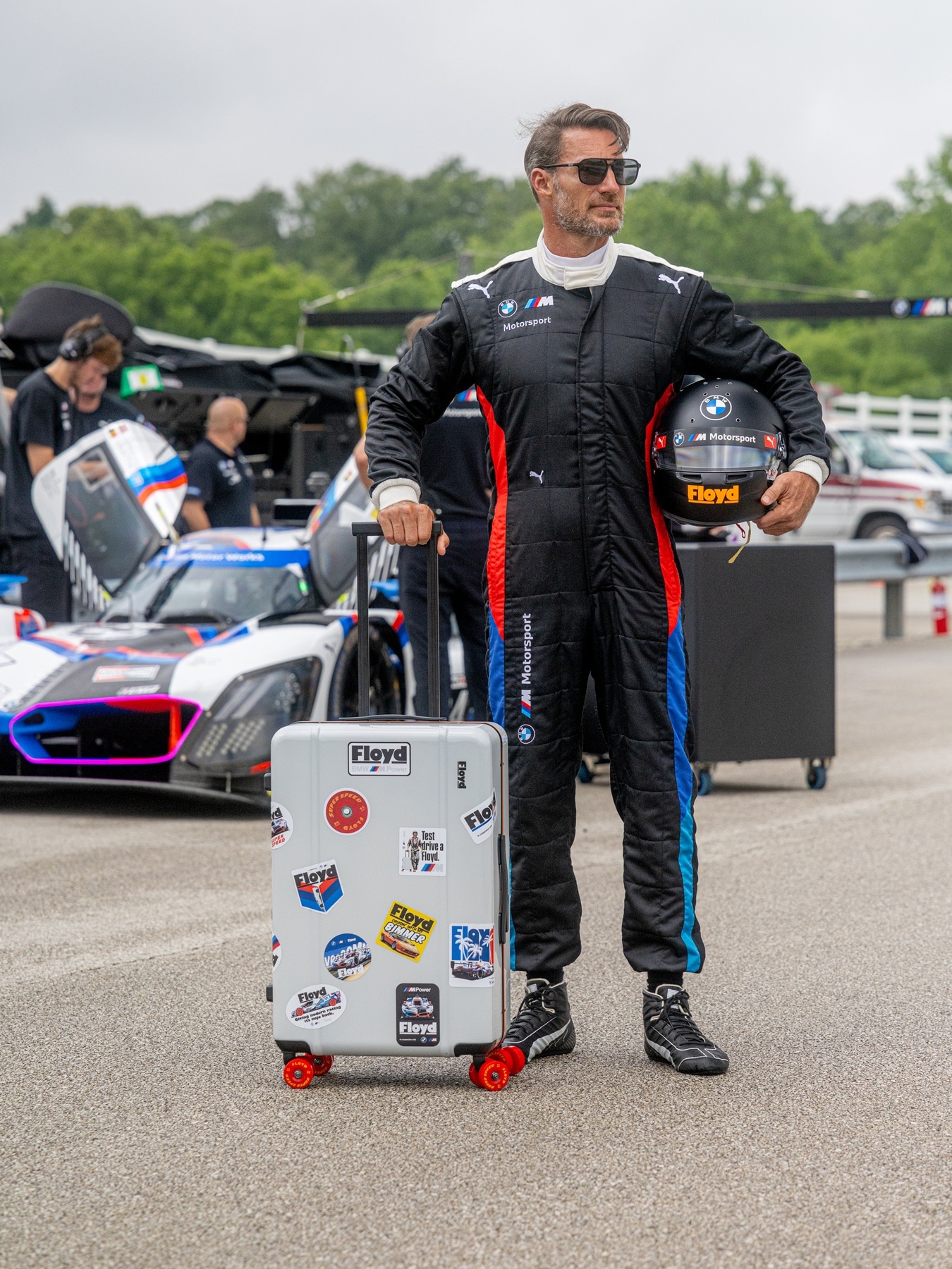 Stuntman mit Floyd-Rollkoffer steht vor Rennfahrzeug BMW M Hybrid V8