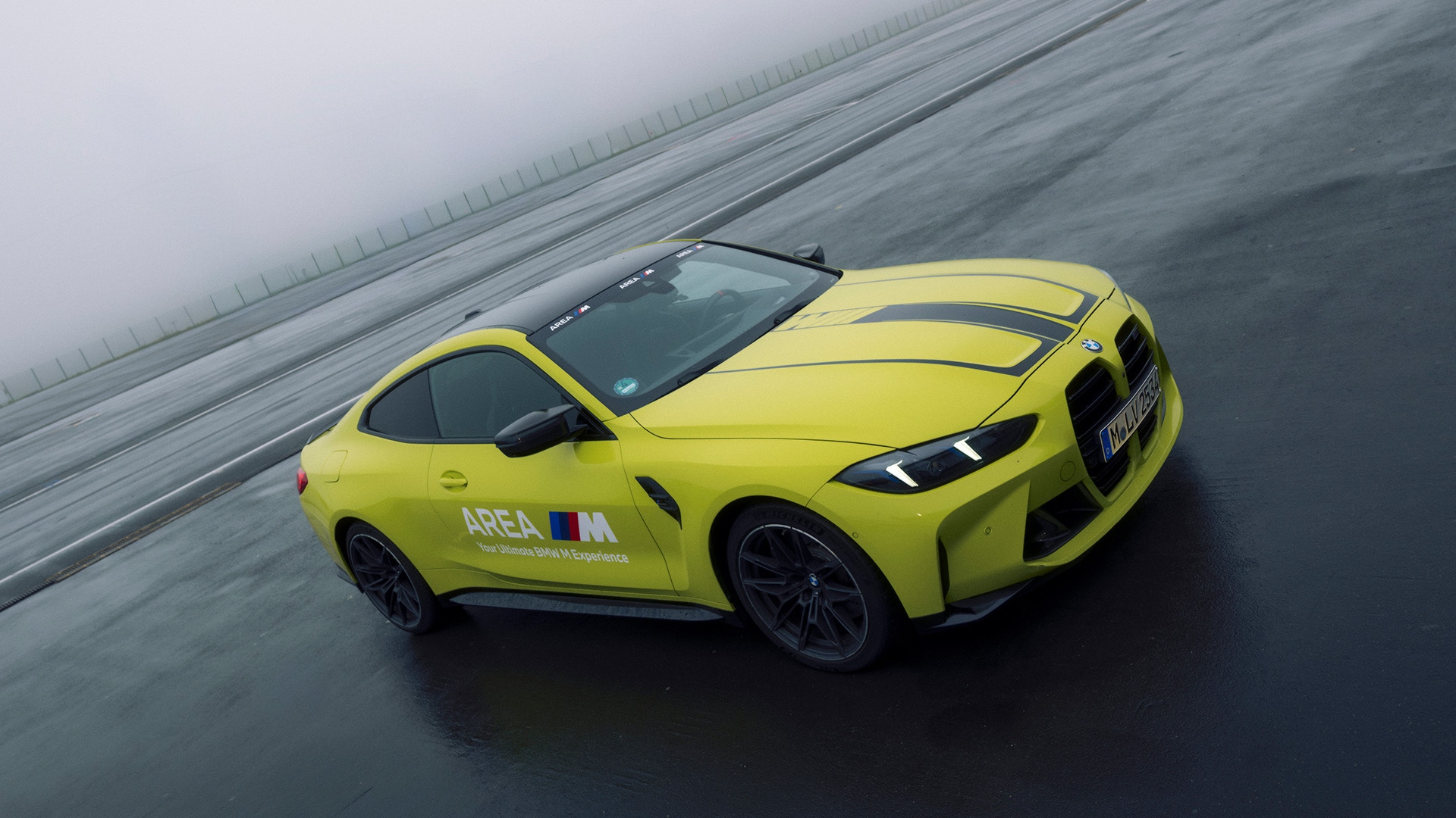 A bright yellow BMW sports car with 'AREA M' branding on its side, positioned on a wet racetrack or driving course. The car is photographed from above on a misty, overcast day with the track surface appearing dark and slick.
